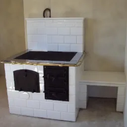 Biały piec kaflowy z żeliwną płytą grzewczą i piekarnikiem, ozdobiony mosiężnymi detalami, z wbudowanym siedziskiem wyłożonym kafelkami.