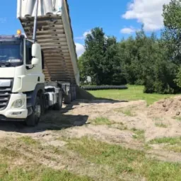 Wyładowujący się z wywrotki piasek na placu budowy, w tle palety z kostką brukową i budynek gospodarczy.