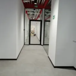 Wnętrze biurowe po remoncie: białe ściany, szara podłoga, czarne listwy przypodłogowe i przeszklone drzwi z widoczną drabiną malarską w oddali.