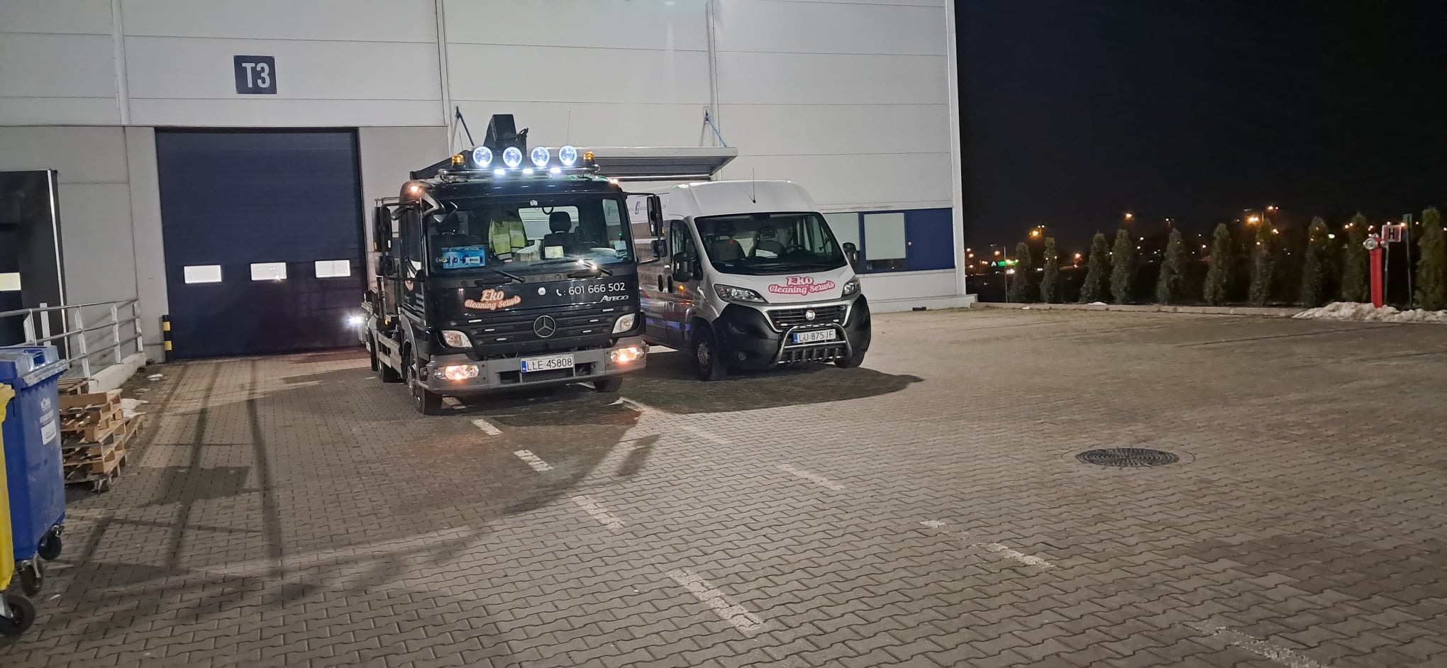 Dwa samochody firmy Eko Cleaning Serwis zaparkowane przed halą magazynową T3 w nocy, widoczny czarny Mercedes z dźwigiem i dodatkowym oświetleniem oraz biały Fiat Ducato.