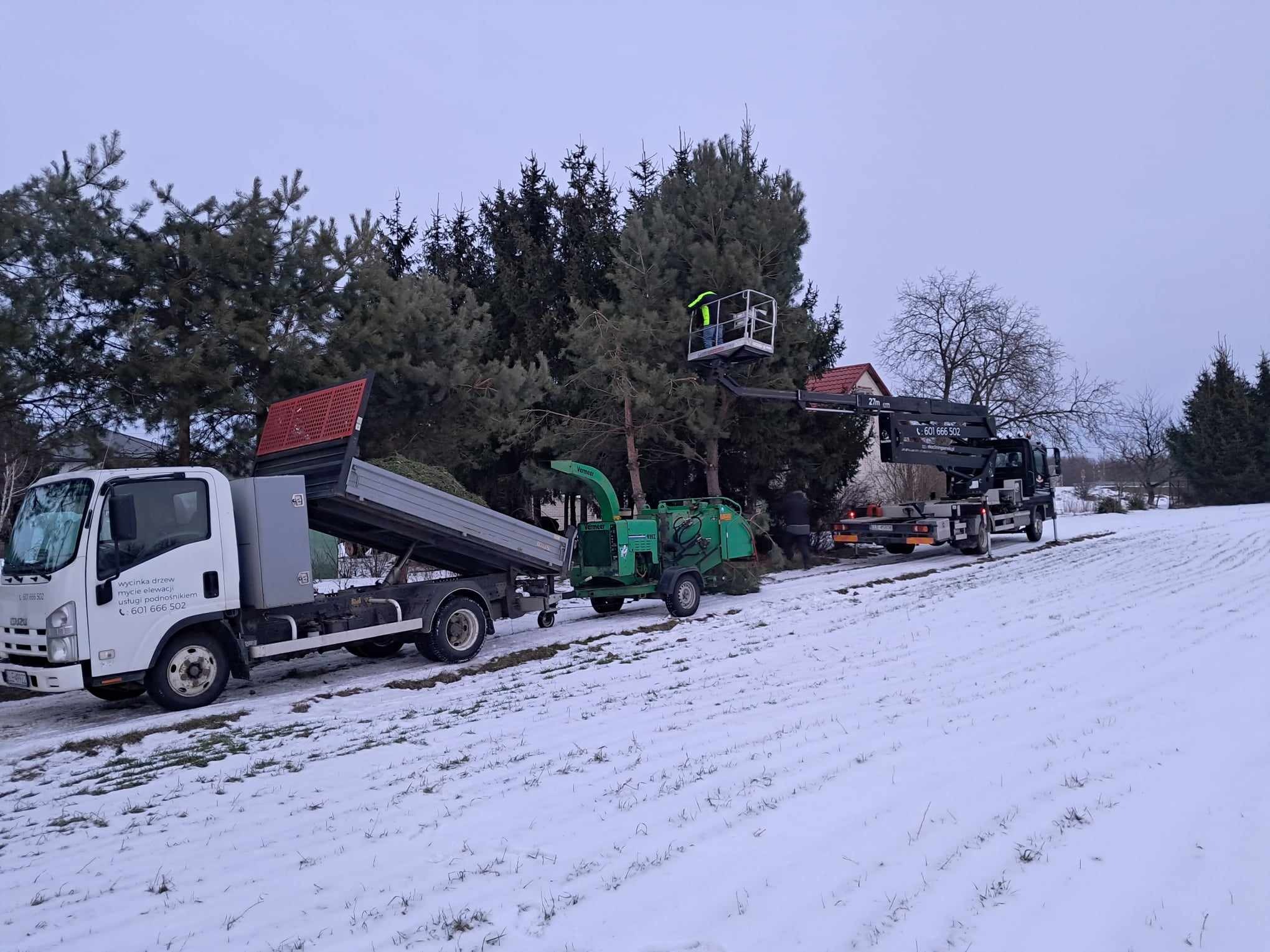 Pracownik w podnośniku koszowym przycina drzewo, gałęzie trafiają do rozdrabniacza, a zrębki do wywrotki na ośnieżonym terenie; na burcie wywrotki widnieje napis 'wycinka drzew, mycie elewacji...