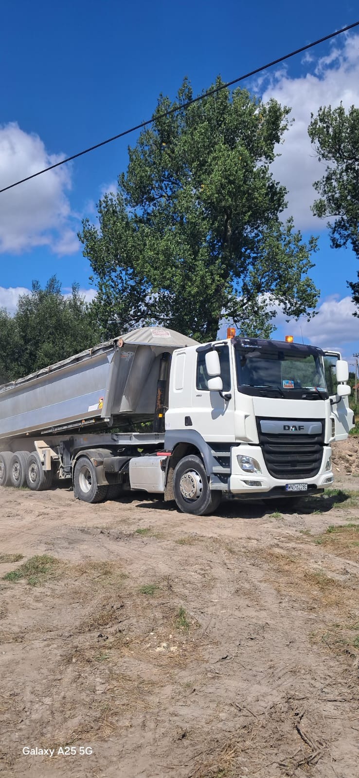 Biała ciężarówka wywrotka DAF z aluminiową naczepą na ziemistym placu budowy, widok z boku, błękitne niebo i drzewa w tle.