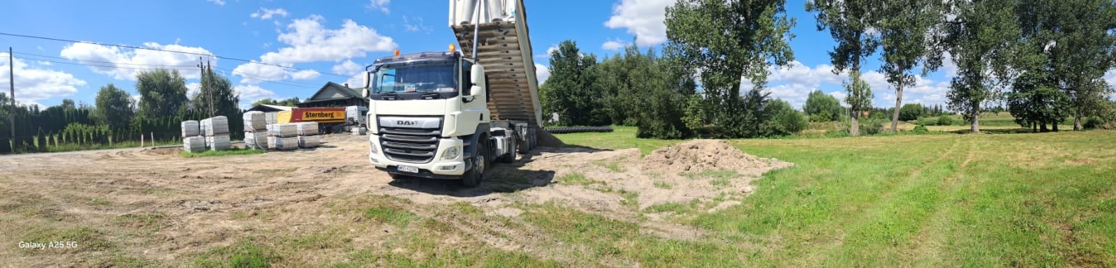 Wyładowujący się z wywrotki piasek na placu budowy, w tle palety z kostką brukową i budynek gospodarczy.