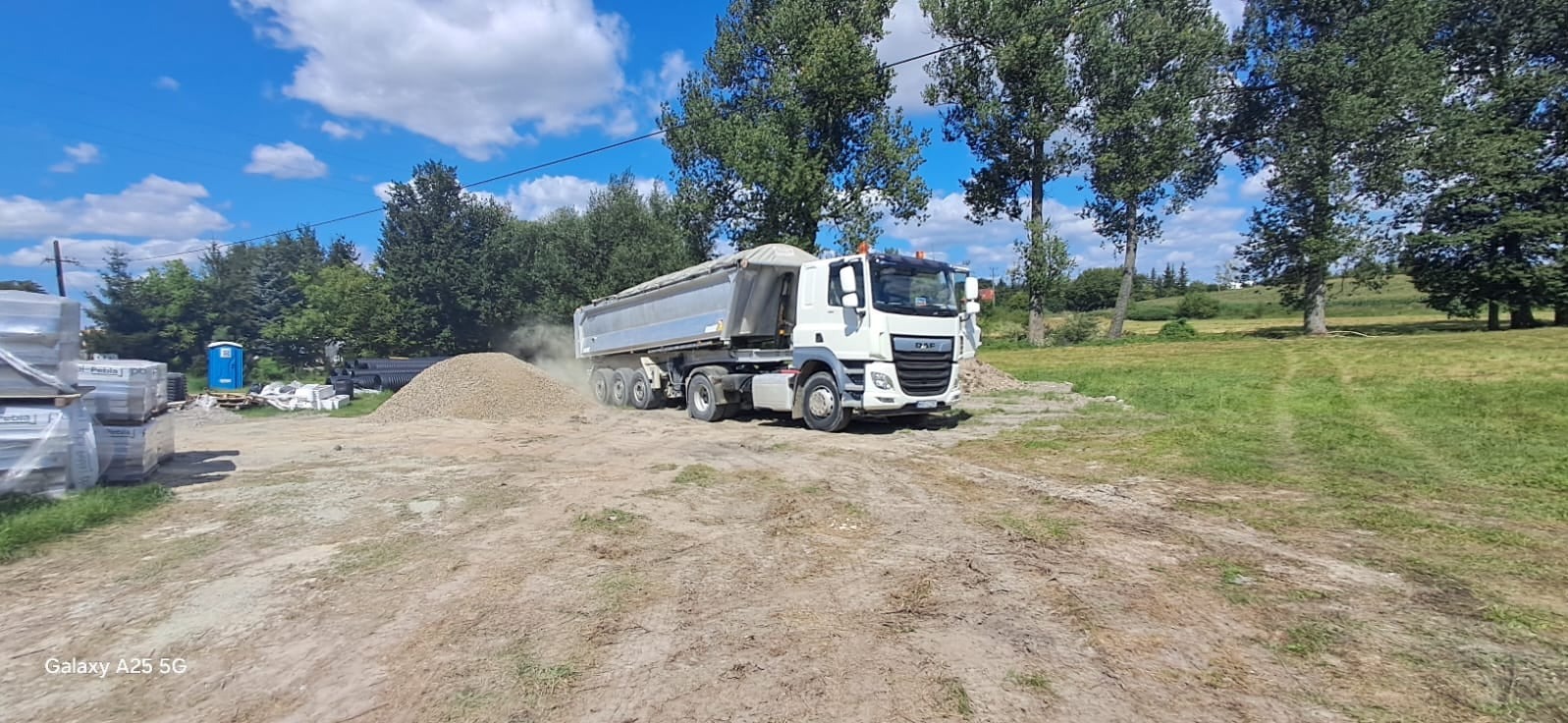 Ciężarówka wyładowuje żwir na placu budowy, w tle widoczne materiały budowlane i przenośna toaleta.