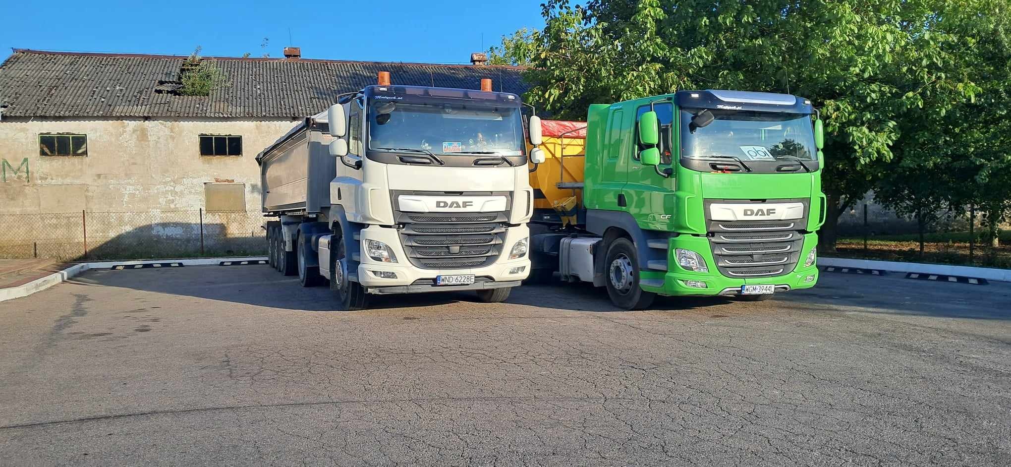 Dwa samochody ciężarowe DAF: biały wywrotka i zielony z żółtym zbiornikiem na posypkę, zaparkowane na placu z budynkiem w tle.