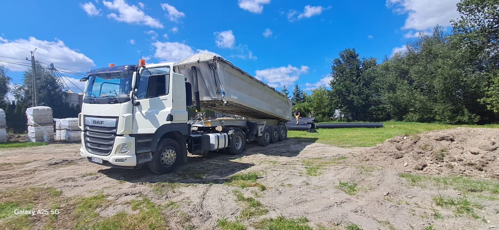 Ciężarówka DAF wywrotka z podniesioną skrzynią ładunkową na placu budowy, widoczne rury drenażowe i stosy kostki brukowej w tle, błękitne niebo z chmurami.