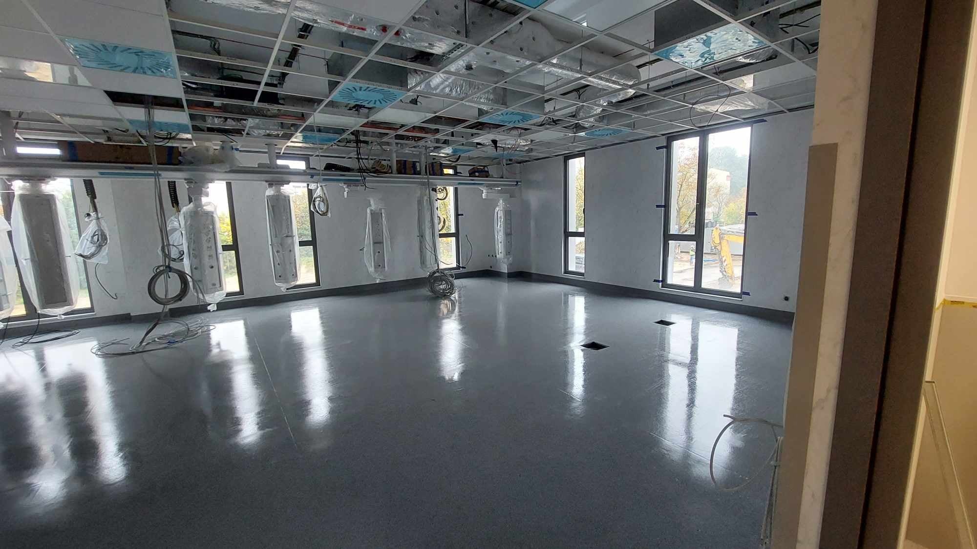 Wnętrze nowego biura z szarą podłogą i białymi ścianami, widoczne instalacje pod sufitem, okna z widokiem na zewnątrz, stan po remoncie.