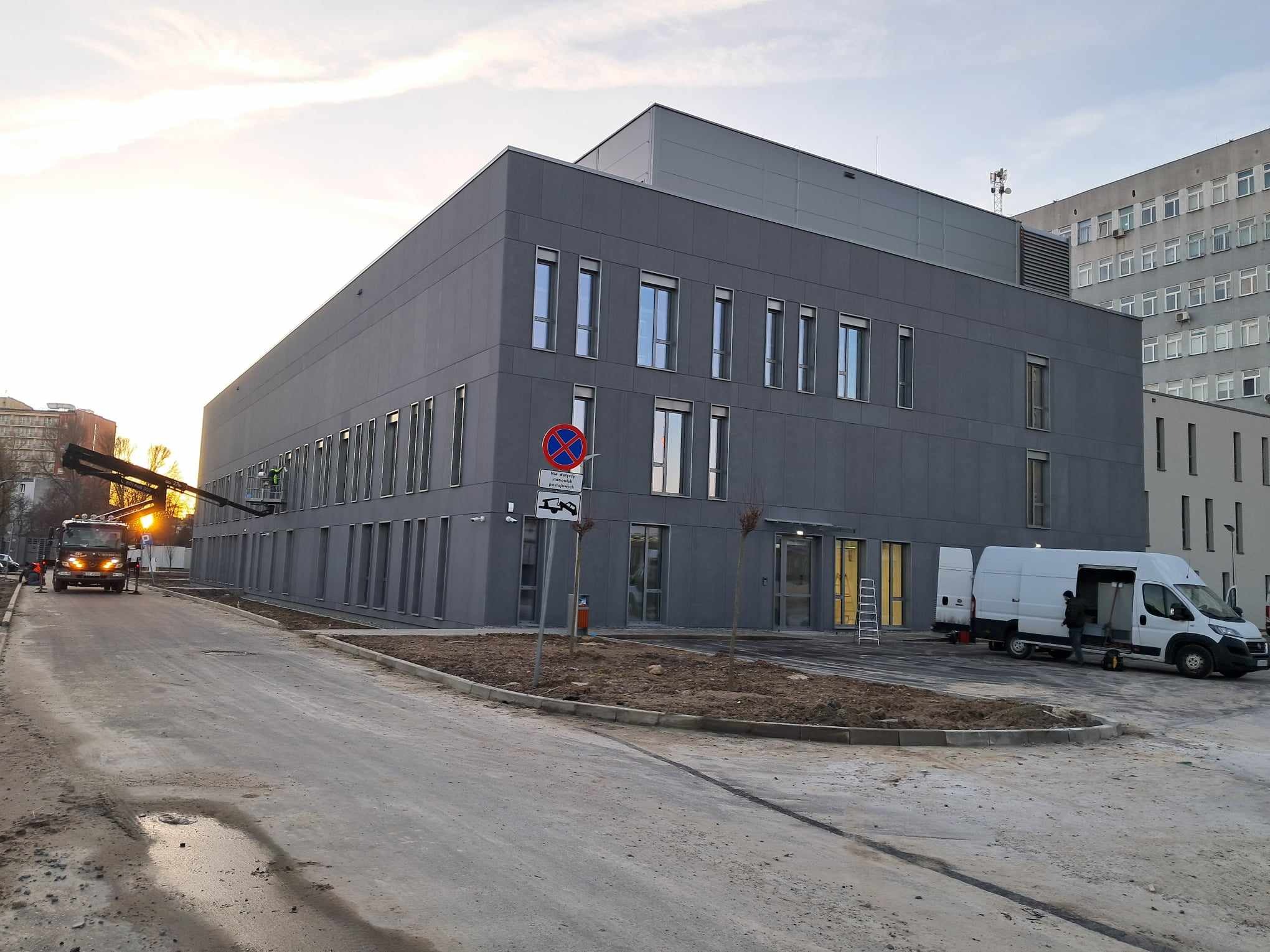 Szara elewacja budynku, myta z podnośnika koszowego, z zaparkowanym busem i widocznym znakiem zakazu postoju