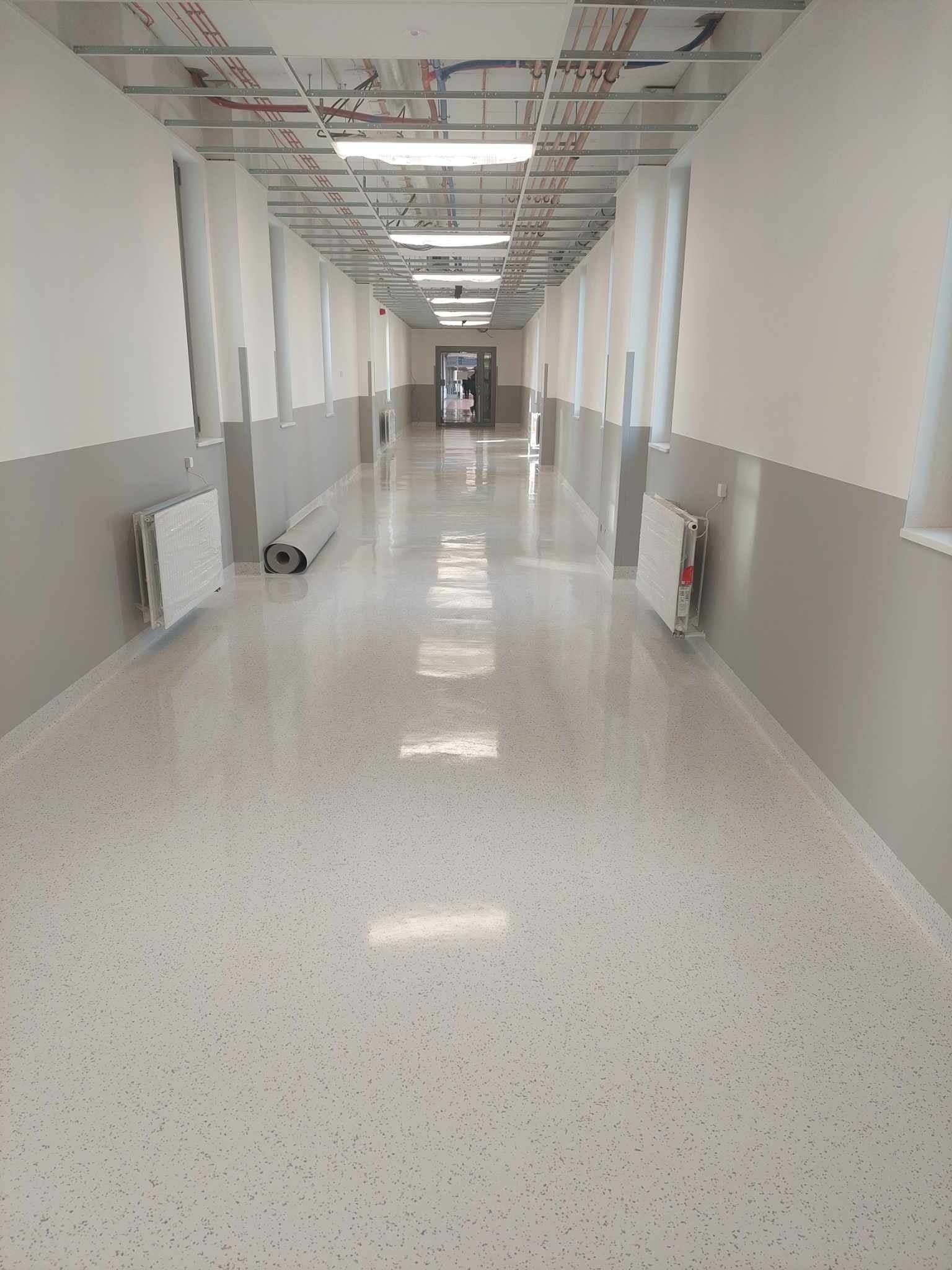 Długi korytarz z połyskującą, jasną podłogą z drobnymi wzorami, świeżo pomalowane ściany w odcieniach szarości i bieli, z grzejnikami w folii ochronnej i rolką materiału pod ścianą.