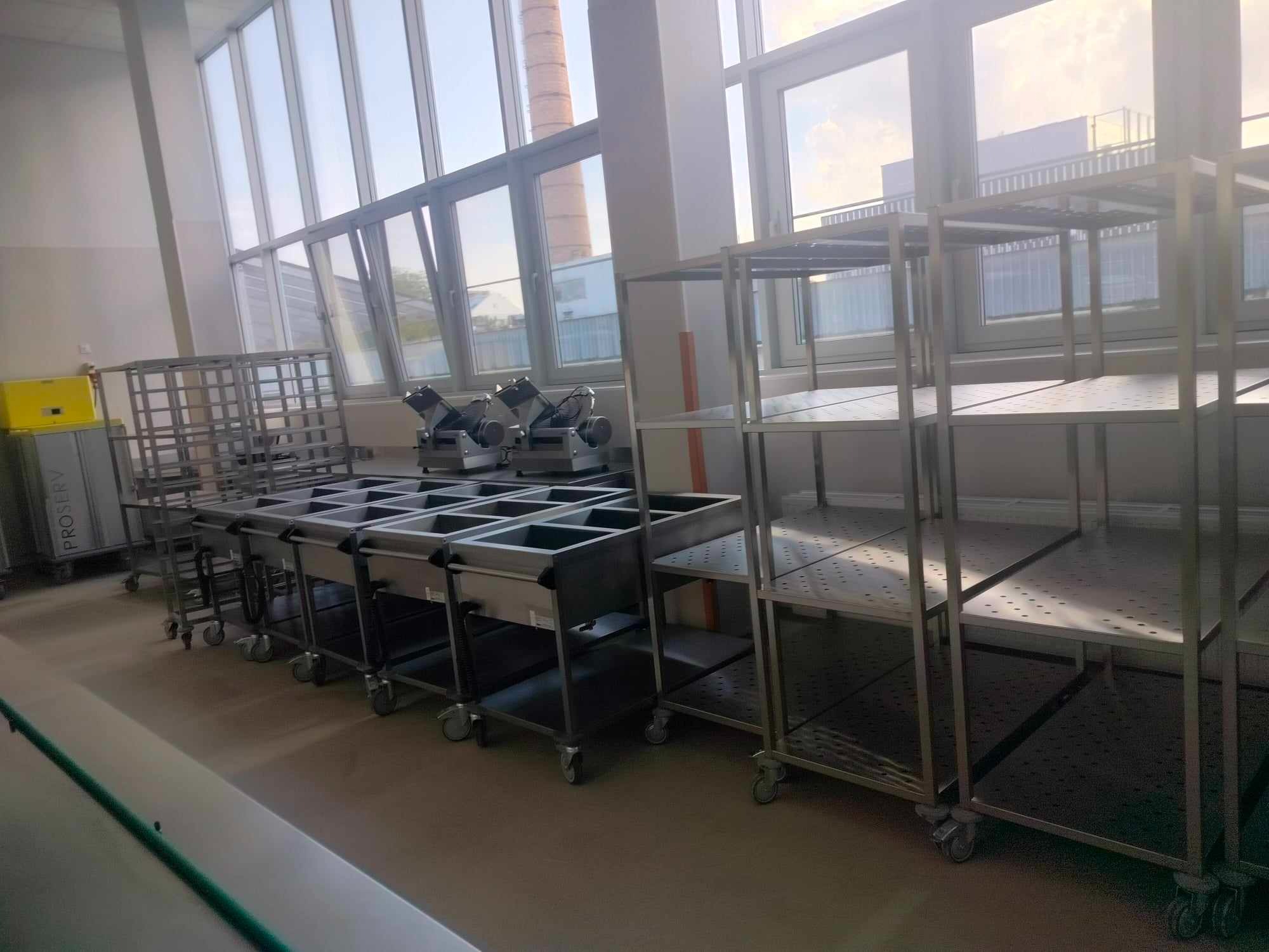 Puste, metalowe regały i wózki gastronomiczne na kółkach w jasnym pomieszczeniu z dużymi oknami.
