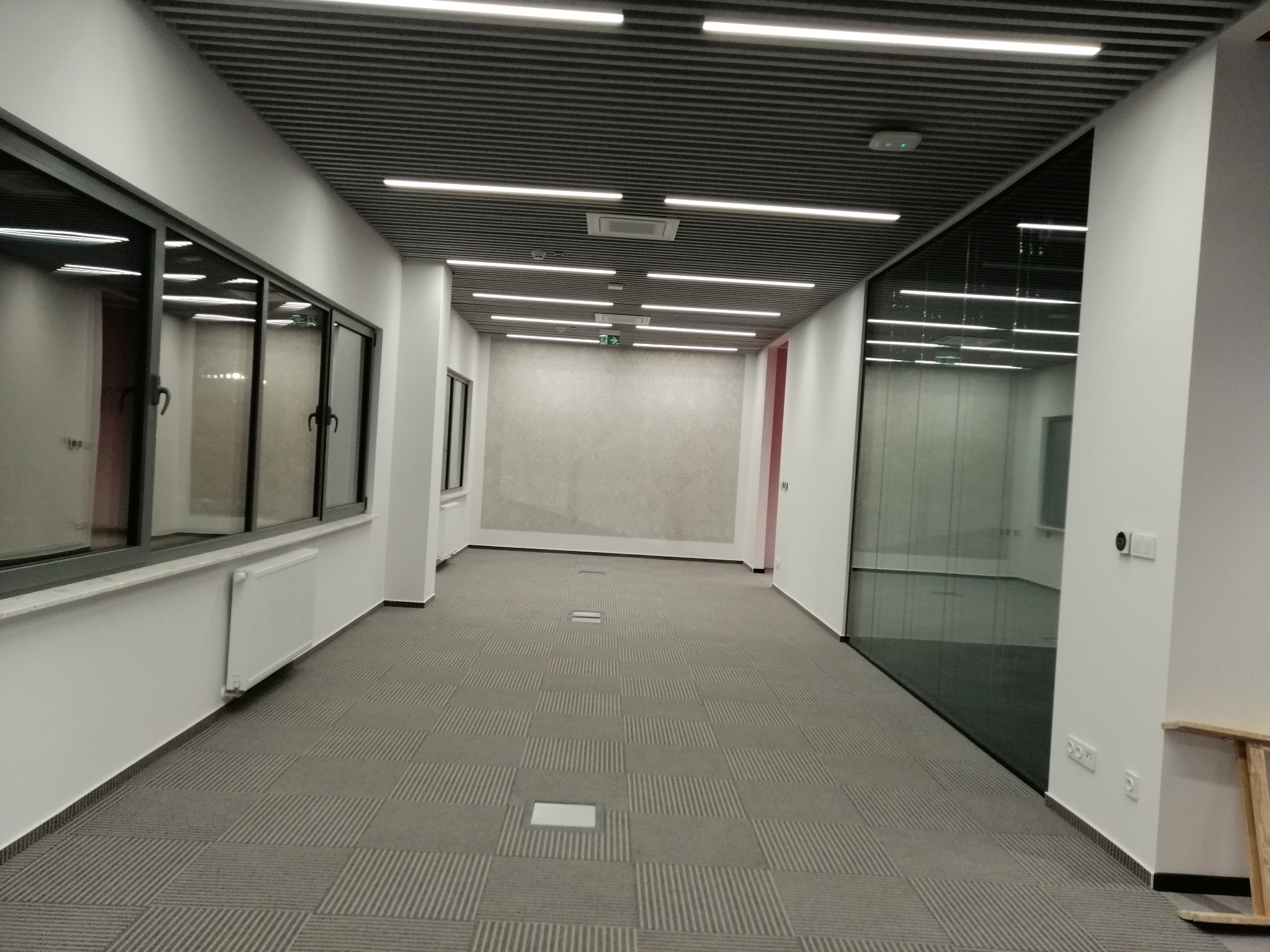 Wnętrze biurowe: Długi korytarz z szarą wykładziną w kwadraty, oświetlenie liniowe na suficie, ściany białe z dużymi oknami i szklanymi panelami, widoczny fragment drabiny.