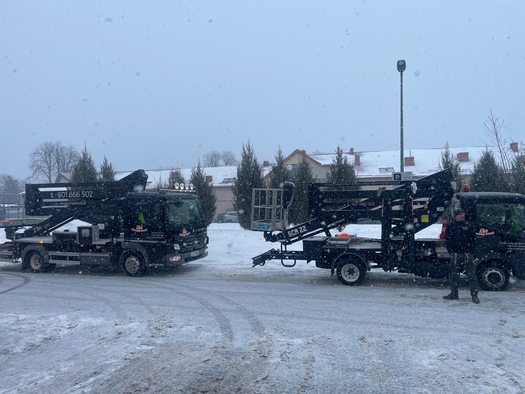 Dwa podnośniki koszowe firmy Eko Cleaning Serwis na ośnieżonym placu podczas opadów śniegu. Widoczne numery kontaktowe i logo firmy na pojazdach.