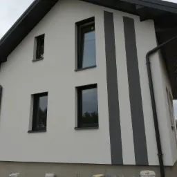 Dwukondygnacyjny dom z jasną elewacją i grafitowymi pasami, czarnymi oknami i rynnami, widok z boku na tle pochmurnego nieba.