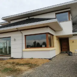 Nowoczesny dom jednorodzinny z płaskim dachem, dużymi oknami z brązowymi ramami i częściowo otynkowaną elewacją w kolorach bieli i jasnej żółci, widoczny fragment posesji z szarym żwirem, samochód...