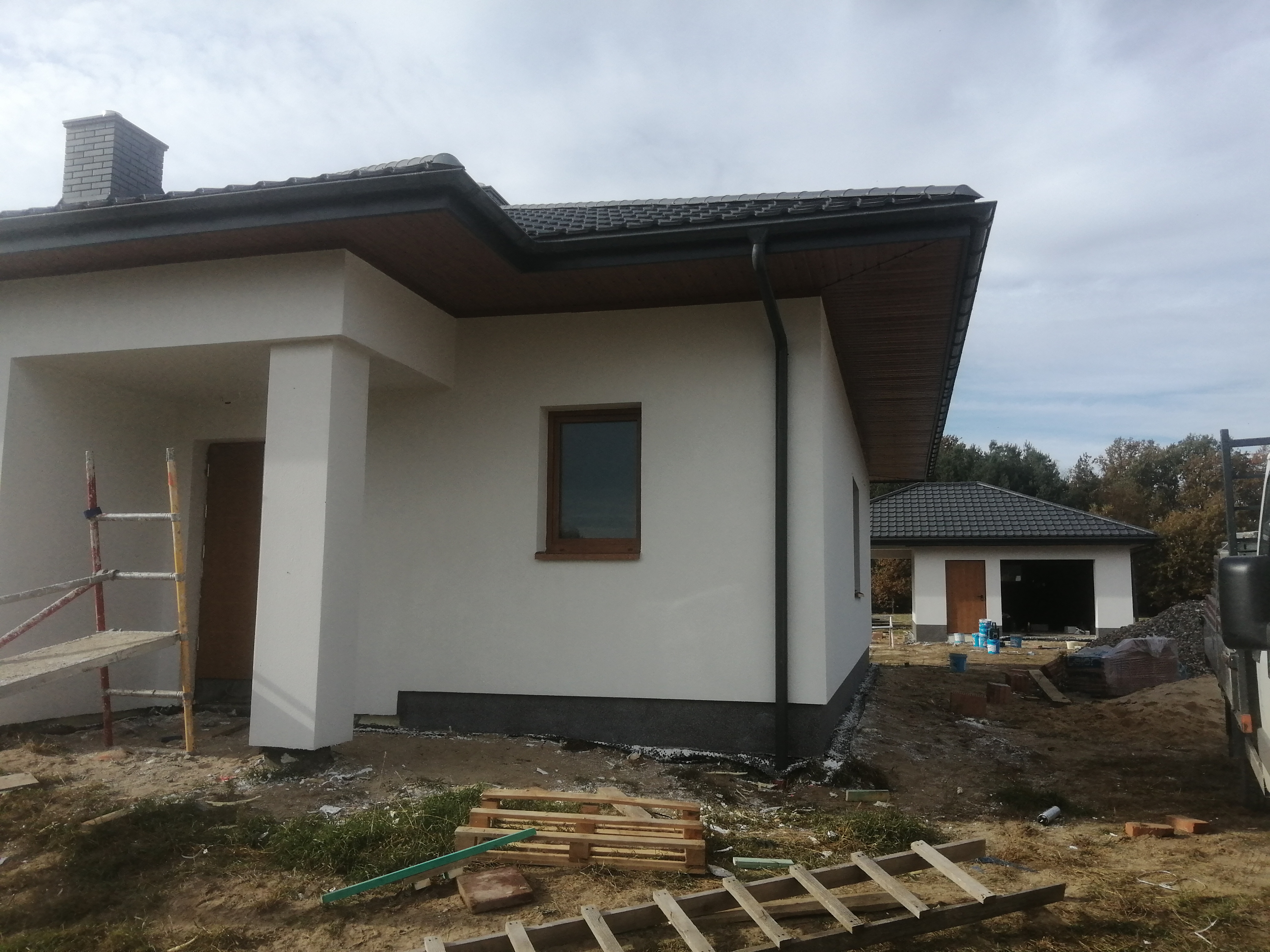 Nowoczesny dom jednorodzinny z białymi ścianami i ciemną dachówką, widoczny fragment elewacji oraz garaż w tle, teren wokół w trakcie prac wykończeniowych, widoczne rusztowanie i materiały budowlane.