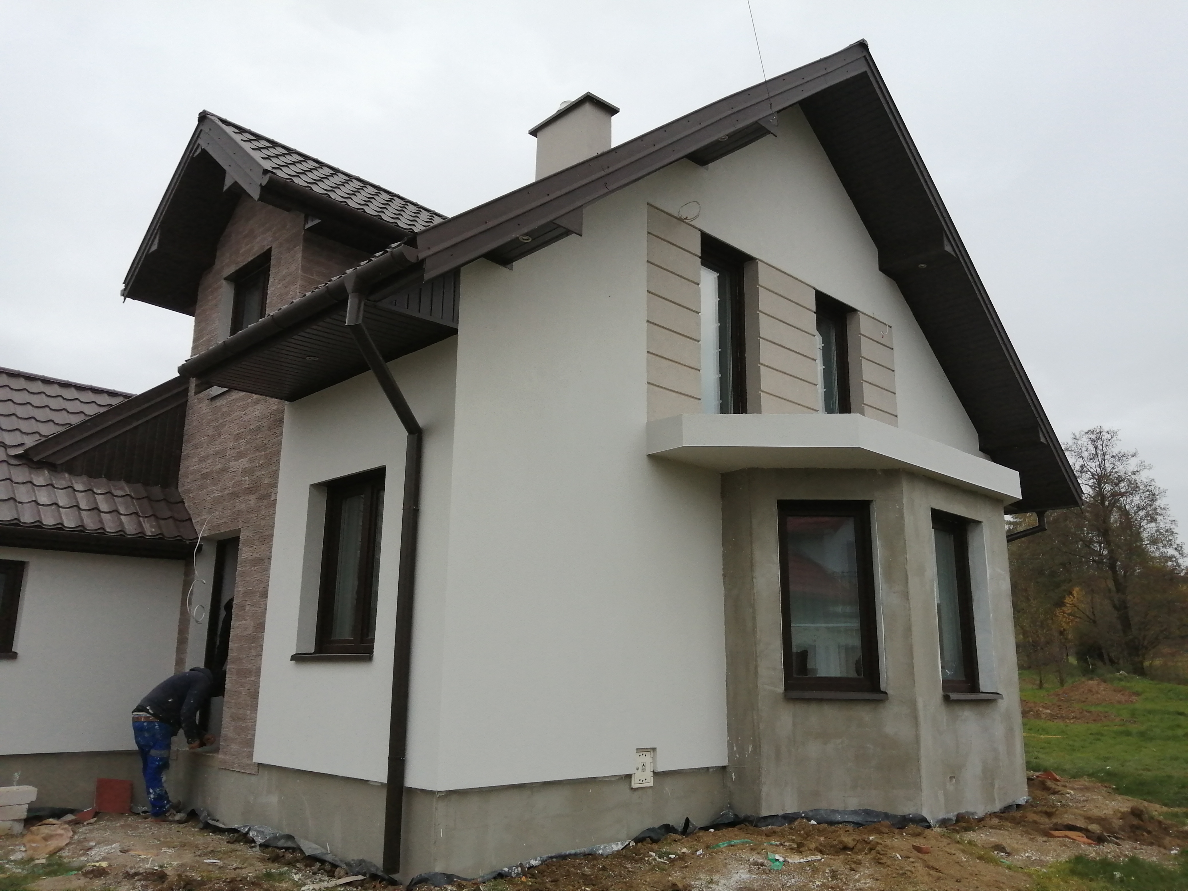 Dwukondygnacyjny dom z białymi i ceglanymi ścianami, brązowym dachem i oknami, w trakcie prac wykończeniowych na zewnątrz, z widocznym pracownikiem w niebieskich spodniach i czarnej bluzie.