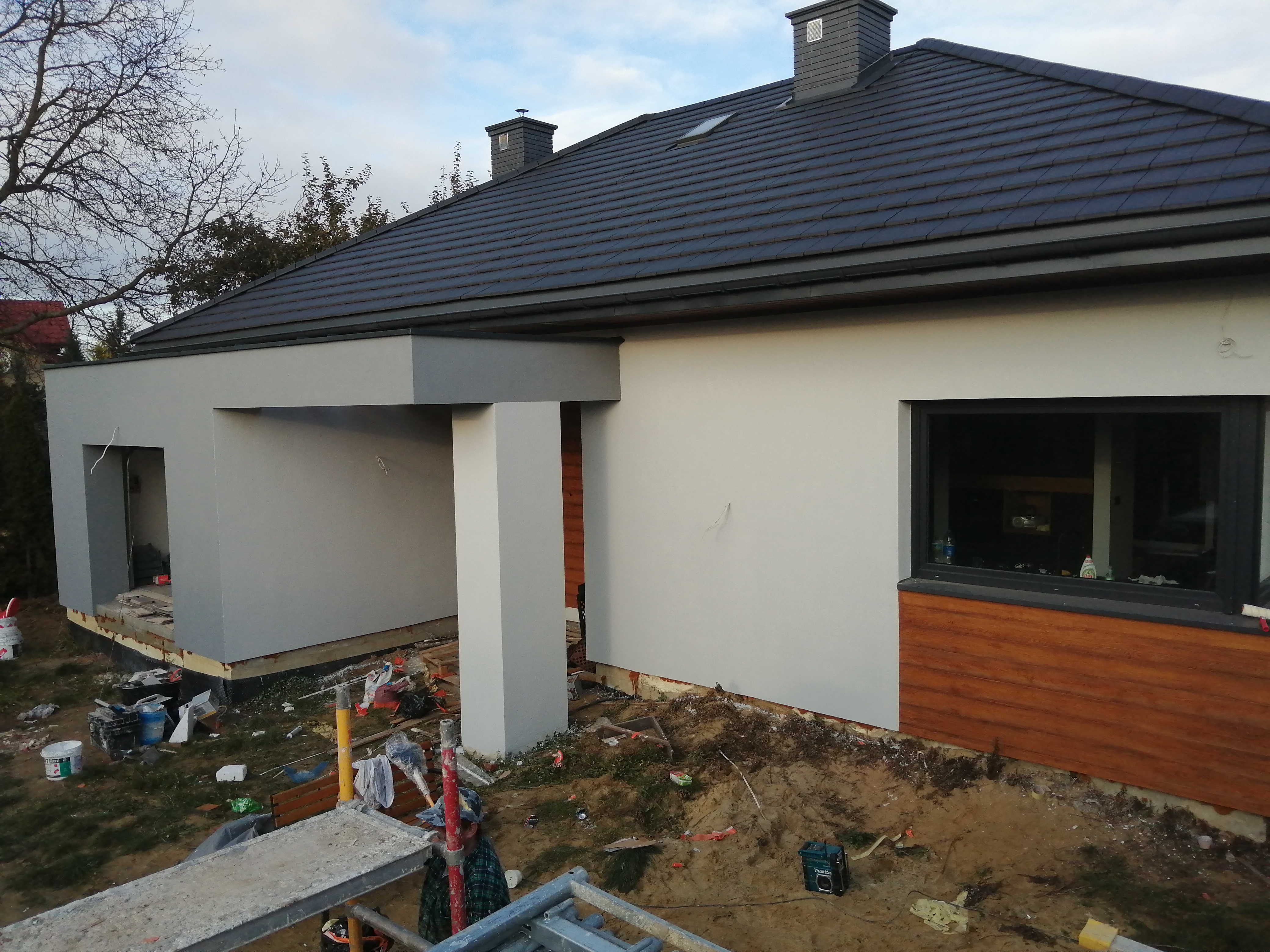 Nowoczesny dom jednorodzinny z szarą elewacją, ciemną dachówką i elementami drewnianej okładziny, widoczne rusztowania i teren w trakcie prac wykończeniowych wokół budynku.