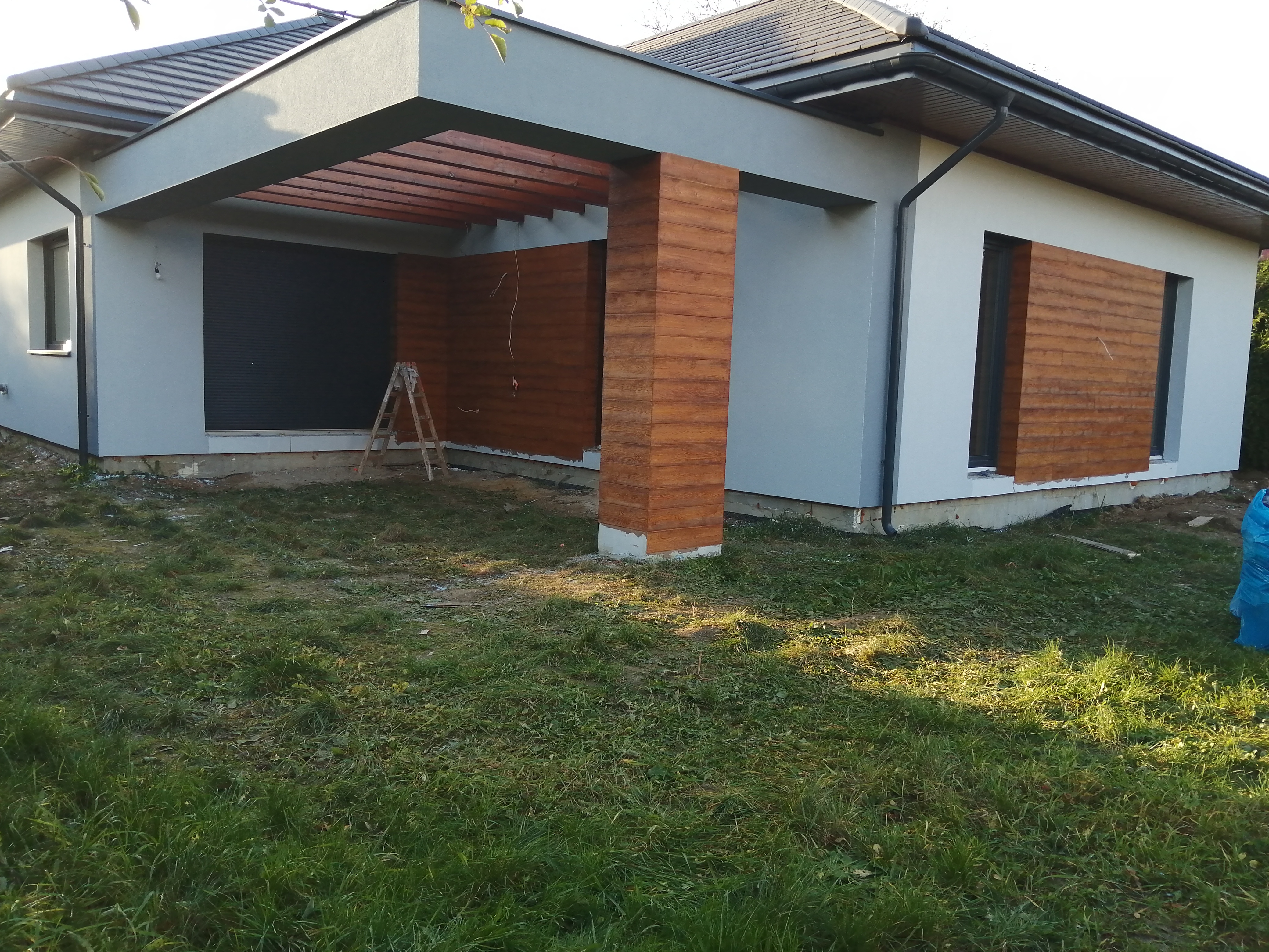 Nowoczesny dom jednorodzinny z szarą elewacją i drewnianymi elementami dekoracyjnymi, widoczne rusztowanie przy ścianie, trawnik w pierwszym planie.