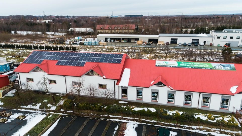 Budynek z czerwonym dachem i panelami słonecznymi na dachu, otoczony zimowym krajobrazem z widocznym banerem reklamowym na dachu.
