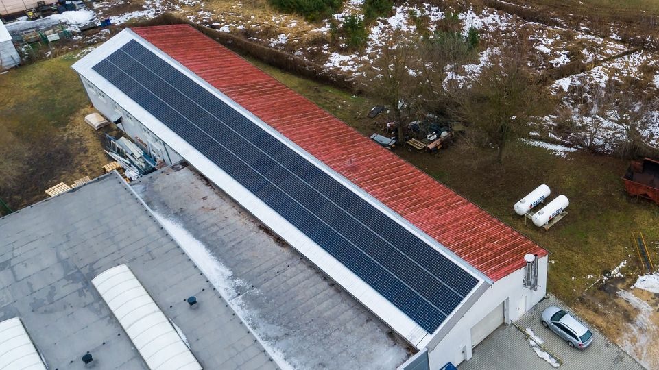 Widok z góry na długi budynek z panelami słonecznymi na dachu. Obok budynków widoczne drzewa, trawa i zaparkowany samochód.