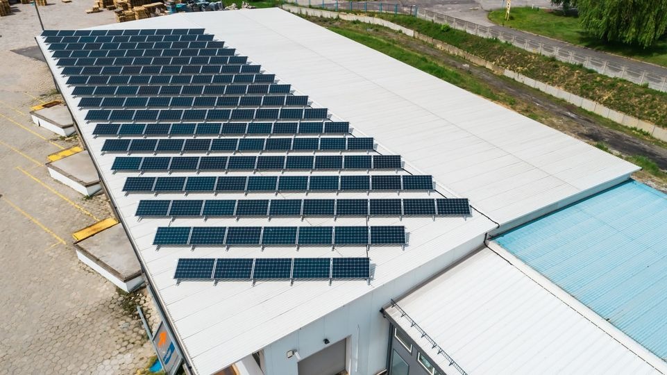 Widok z góry na dużą instalację paneli słonecznych na dachu hali magazynowej z rampami załadunkowymi i betonowym placem.