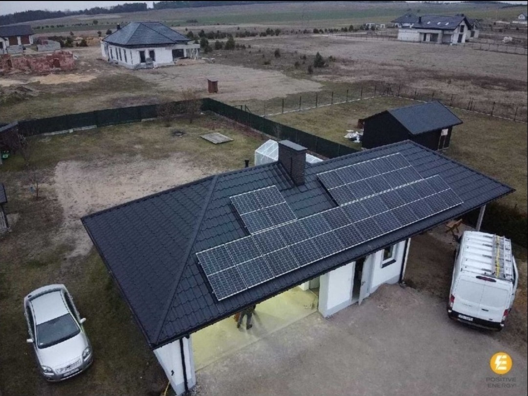Widok z drona na dom z ciemną dachówką i zamontowanymi panelami fotowoltaicznymi; obok zaparkowany srebrny samochód osobowy i biały bus z drabiną na dachu.