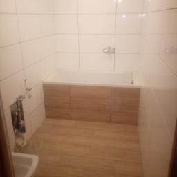 Andrzej Graca - Wykończona łazienka z białymi płytkami ściennymi, drewnopodobnymi płytkami podłogowymi i zabudową wanny z paneli drewnopodobnych; widoczne niezamaskowane rury instalacyjne.