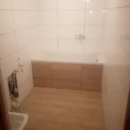 Wykończona łazienka z białymi płytkami ściennymi, drewnopodobnymi płytkami podłogowymi i zabudową wanny z paneli drewnopodobnych; widoczne niezamaskowane rury instalacyjne.