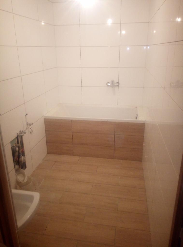 Wykończona łazienka z białymi płytkami ściennymi, drewnopodobnymi płytkami podłogowymi i zabudową wanny z paneli drewnopodobnych; widoczne niezamaskowane rury instalacyjne.