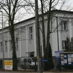 Dwupiętrowy budynek pomalowany na szaro z białymi akcentami, widoczny zza drzew bez liści i ogrodzenia, z tablicą ogłoszeń i zaparkowanym vanem w tle, sfotografowany w pochmurny dzień.