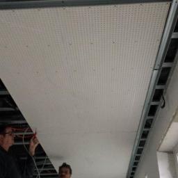 Sebastian wojewoda - Montaż perforowanej płyty sufitowej na metalowej konstrukcji przez dwóch mężczyzn, jeden na drabinie, widoczne narzędzia i przewody.