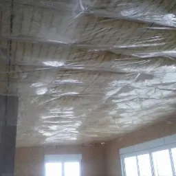 Izolacja poddasza z folią ochronną, widoczne belki konstrukcyjne i okna w ścianie szczytowej.