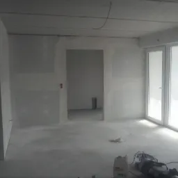Surowe wnętrze w trakcie wykańczania: ściany przygotowane pod malowanie, widoczne ślady szpachlowania, podłoga z wylewką, okna z folią ochronną, narzędzia i materiały budowlane na podłodze.