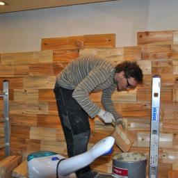 Sebastian wojewoda - Mężczyzna w okularach i rękawicach roboczych nakłada klej na drewnianą płytkę przed przyklejeniem jej do ściany z mozaiką z drewnianych elementów. Obok stoi drabina i poziomica Krause.