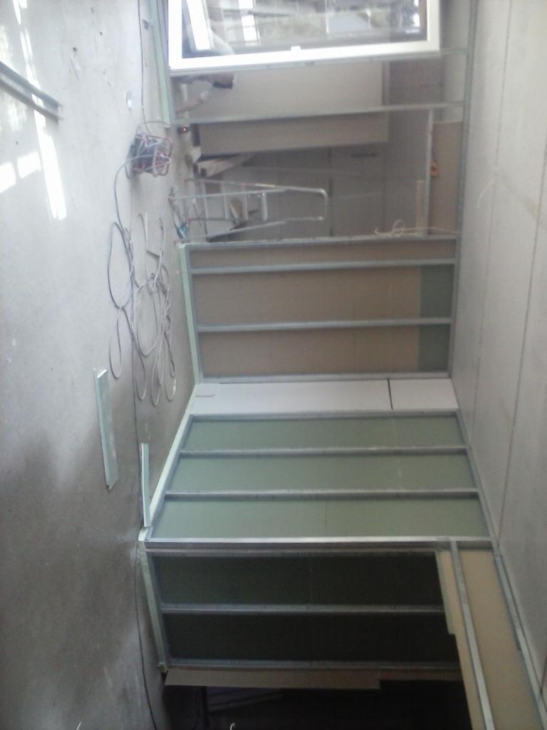 Wnętrze w trakcie prac wykończeniowych: metalowa konstrukcja pod zabudowę, częściowo wypełniona zielonymi płytami, widoczne kable i drabina w tle, surowe ściany.