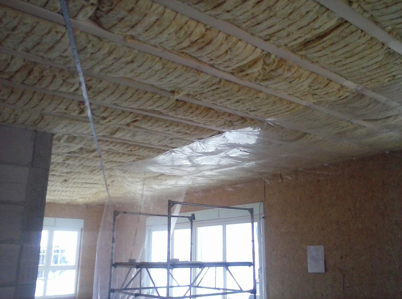 Ocieplenie poddasza wełną mineralną między drewnianymi belkami, zabezpieczone folią paroizolacyjną, widoczne rusztowanie i okno.