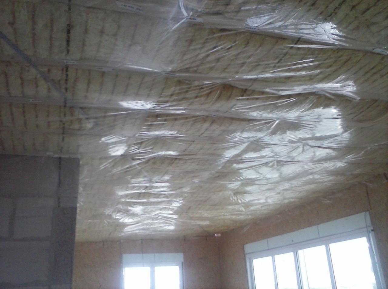 Izolacja poddasza z folią ochronną, widoczne belki konstrukcyjne i okna w ścianie szczytowej.