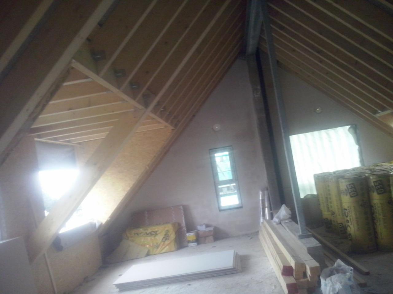 Poddasze w trakcie adaptacji: widoczne drewniane elementy konstrukcyjne dachu, ocieplenie w rolkach, płyty gipsowo-kartonowe i fragment otynkowanej ściany z oknem.