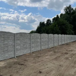 Długi, szary mur z prefabrykowanych elementów o fakturze kamienia, oddzielający teren z drzewami i błękitnym niebem od piaszczystej drogi z wyraźnymi śladami opon.