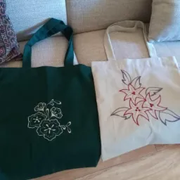 Dwie torby bawełniane z haftowanymi motywami kwiatowymi: ciemnozielona z białym haftem i kremowa z czerwono-szarym haftem, leżące na jasnej sofie i drewnianej podłodze.