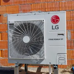 Zewnętrzna jednostka pompy ciepła LG Therma V zamontowana na metalowej konstrukcji przy ścianie z cegły ceramicznej, widoczne przyłącza i cień na murze.