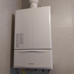 ENERGO-MAX Mariusz Gańczak - Biały, wiszący piec gazowy Immergas z żółtym przewodem gazowym na tle jasnych, kwadratowych płytek. Widoczne zawory i rura odprowadzająca spaliny.