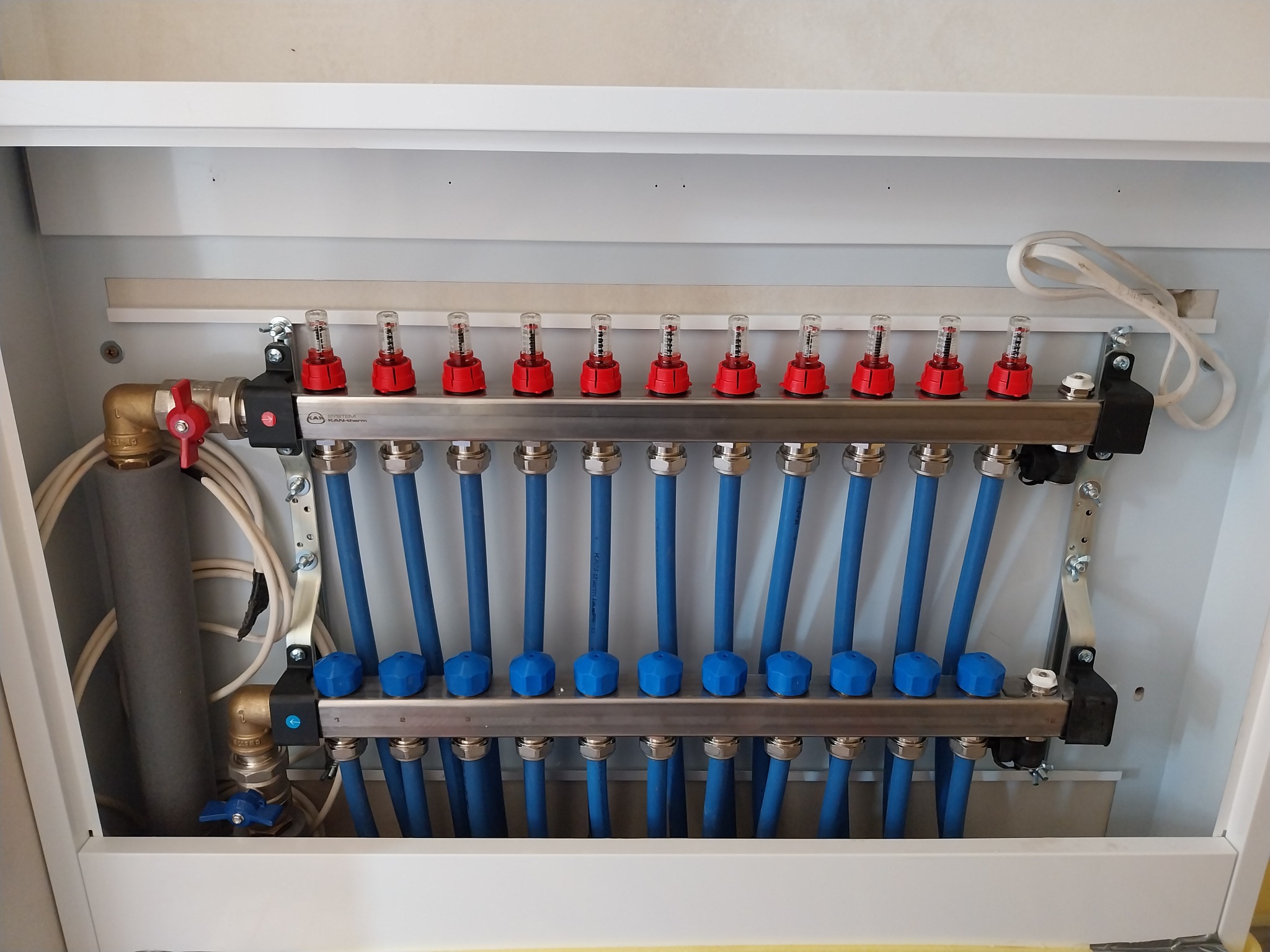 Rozdzielacz ogrzewania podłogowego w białej szafce, z widocznymi niebieskimi rurami, czerwonymi przepływomierzami i niebieskimi zaworami regulacyjnymi, zamontowany na ścianie wewnątrz budynku.