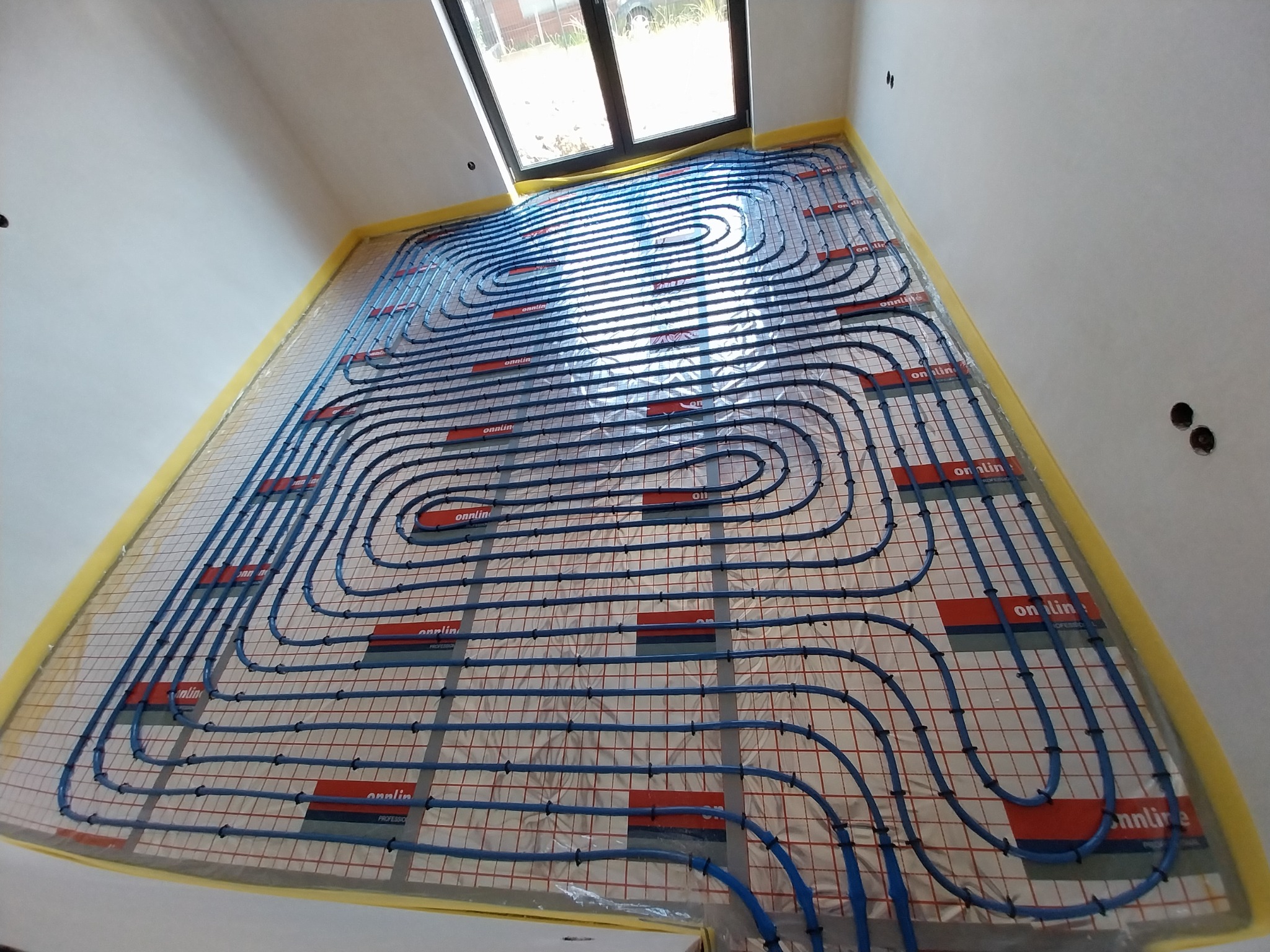 Widok z góry na rozłożoną instalację ogrzewania podłogowego z niebieskich rurek zamocowanych na siatce, zabezpieczoną żółtą taśmą malarską przy ścianach, z oknem dachowym w tle.