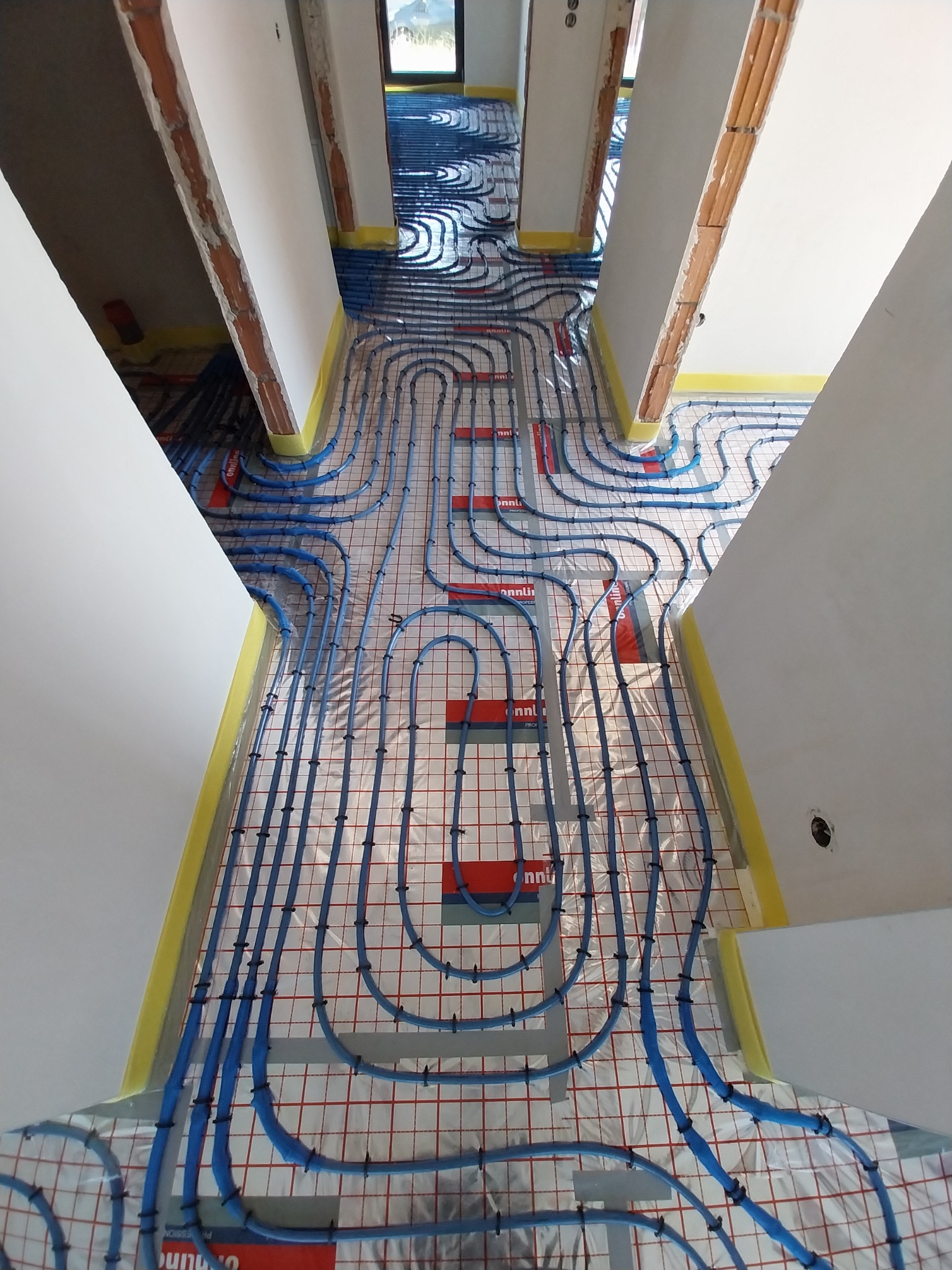 Widok z góry na rozłożoną instalację ogrzewania podłogowego z niebieskich rurek, przymocowanych czarnymi klipsami do folii z czerwoną siatką, w surowym wnętrzu z widocznymi fragmentami cegieł...