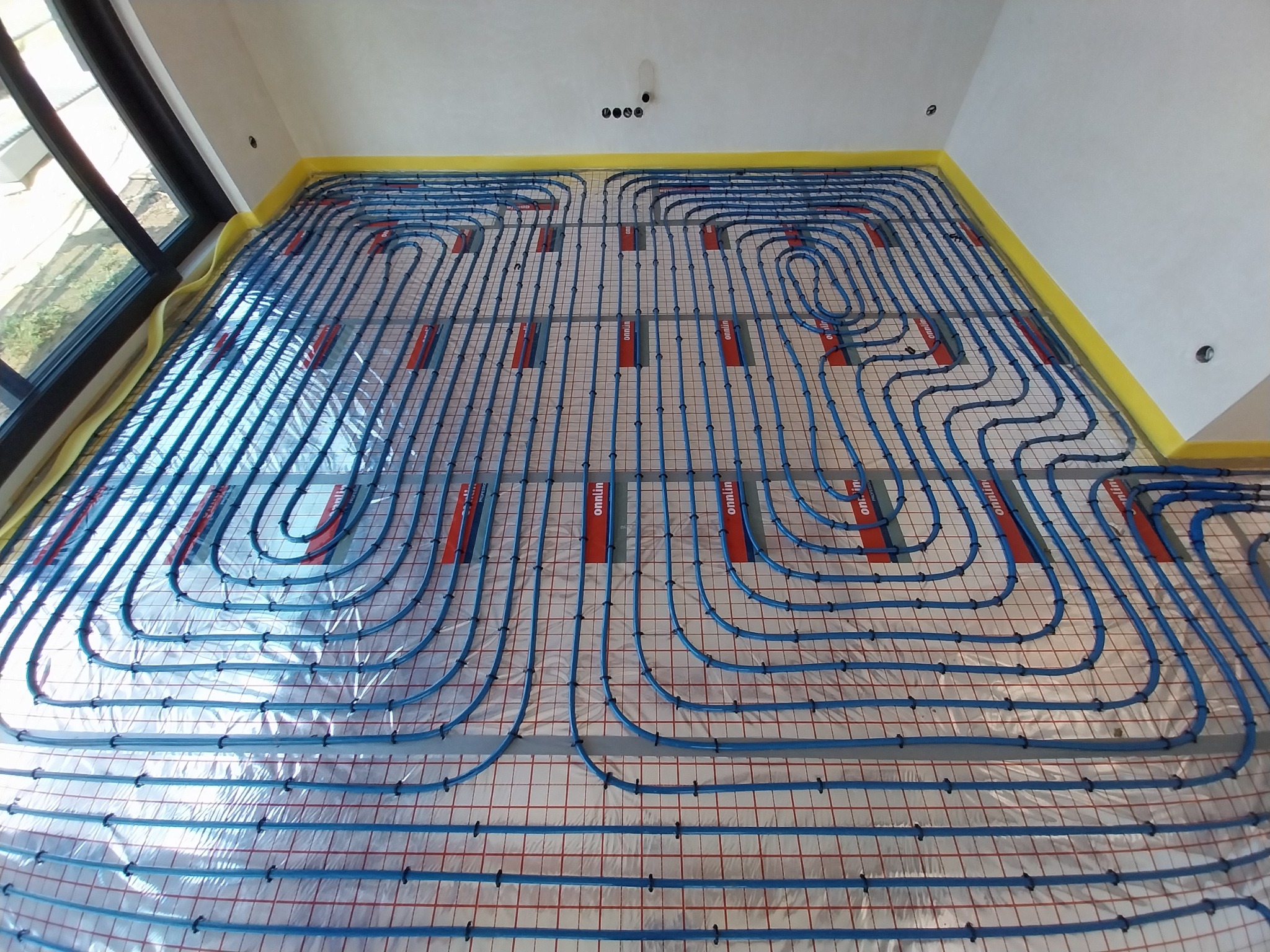 Niebieskie rury ogrzewania podłogowego ułożone na folii z siatką, widok z góry, przygotowane do zalania wylewką, żółta taśma zabezpieczająca ściany.