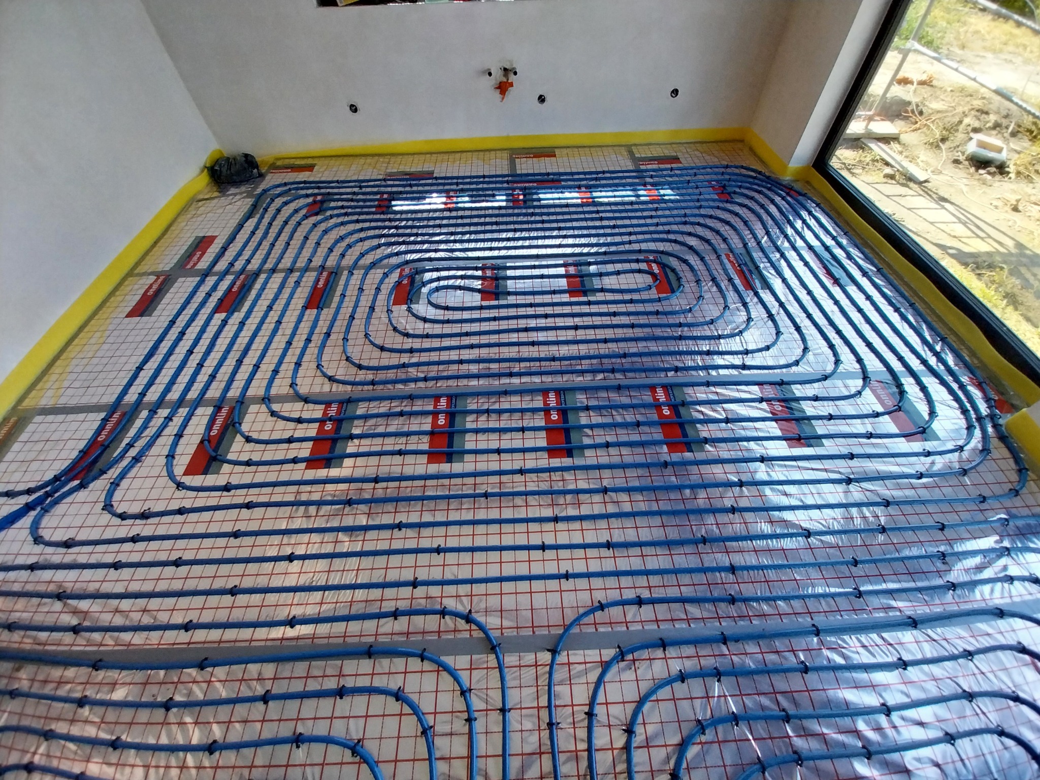 Rozłożone rury ogrzewania podłogowego w nowym budynku, zabezpieczone żółtą taśmą na obwodzie, widoczne mocowania i siatka montażowa.