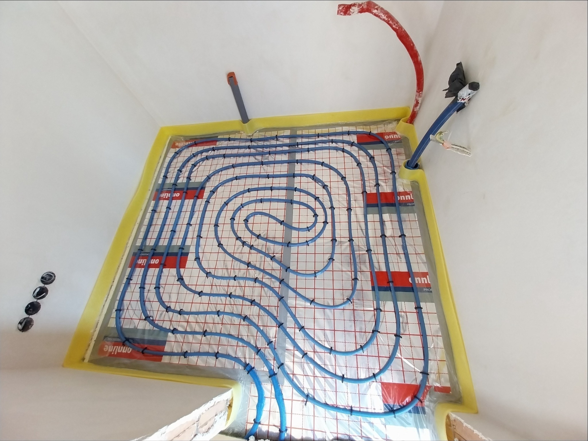 Instalacja podłogowego ogrzewania wodnego z niebieskimi rurami układanymi na macie z czerwoną siatką, zabezpieczona żółtą taśmą malarską przy ścianach, widoczne przyłącza rur.