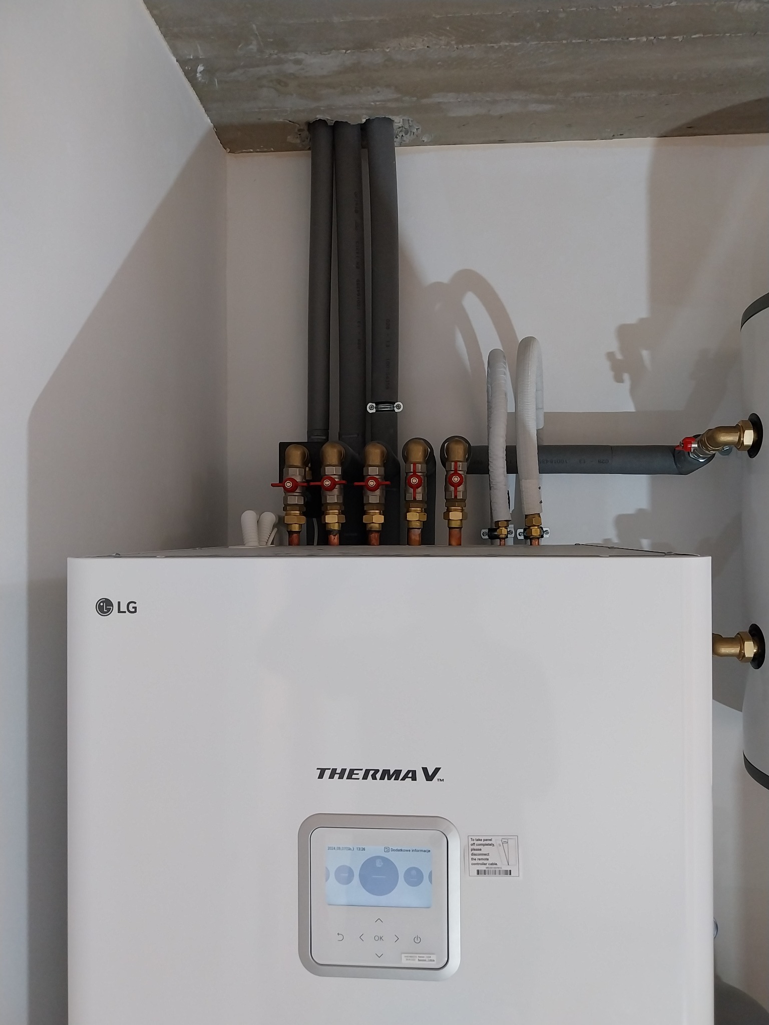 Biała pompa ciepła LG Therma V zamontowana w rogu pomieszczenia, widoczne rury izolowane termicznie i zawory kulowe z czerwonymi rączkami, surowy betonowy sufit.