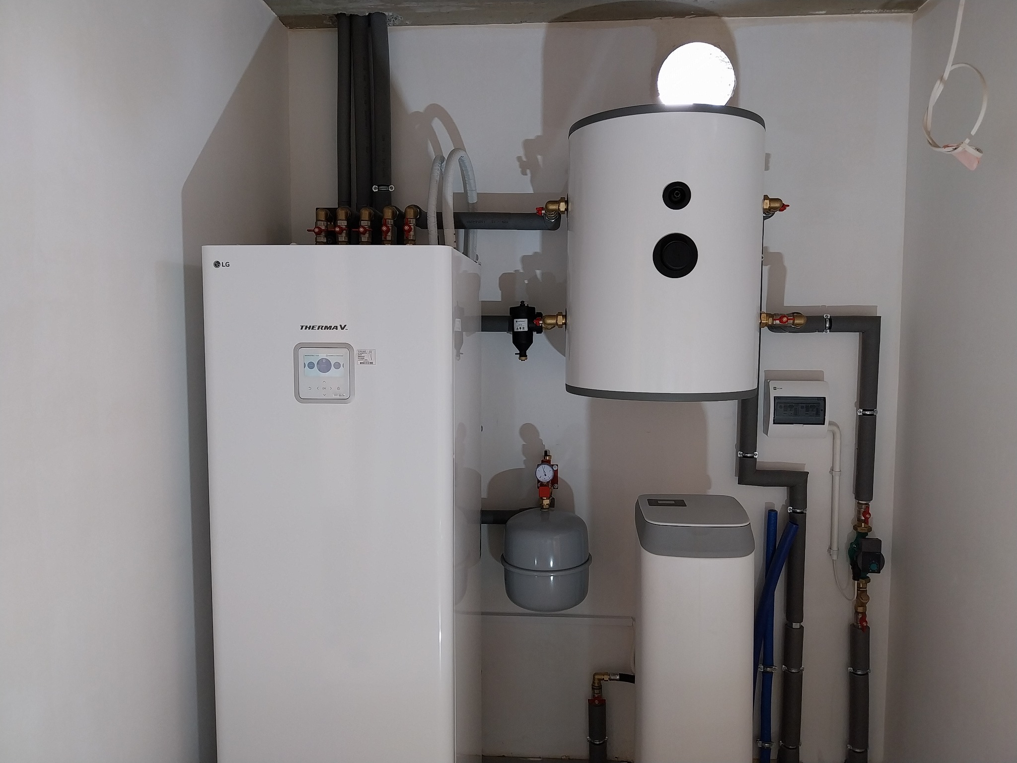 Biała pompa ciepła LG Therma V z podłączonymi rurami instalacji grzewczej i zbiornikiem buforowym w narożniku pomieszczenia.